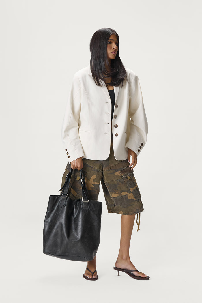 Woman holding a black leather bag wearing a white blazer and camouflage pants on a white background