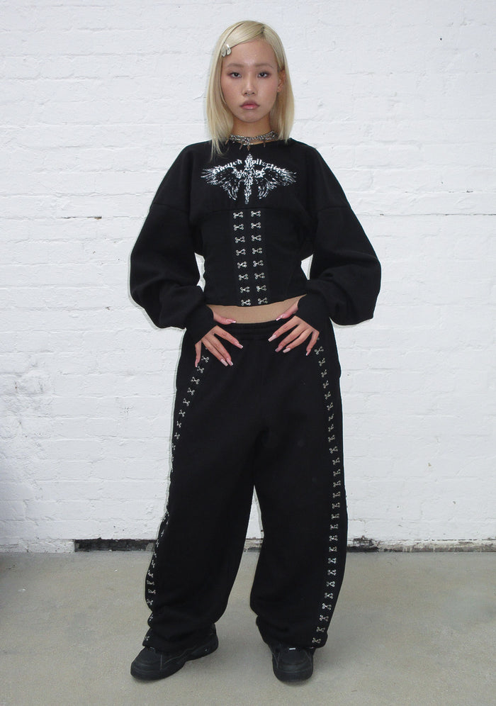 Model wearing a black hoodie featuring a corset waist and decorative detailing, styled with matching black pants.